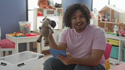 Fototapeta premium Man holds elephant puppet and tablet, points puppet with hand while smiling and seated in building; playful teaching.