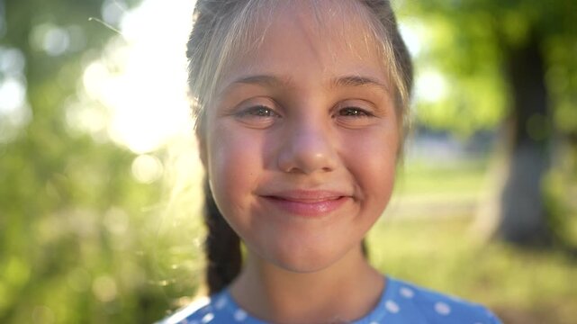 the little girl smiles. concept of a happy childhood and loving family. girl schoolgirl looks at the camera and lifestyle smiles widely, face close-up, park and sun glare in the background