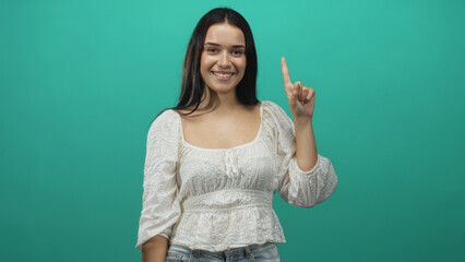 Fototapeta premium Woman smiling and pointing index finger upward in studio, wearing white blouse and jeans against teal backdrop; confidence clarity.