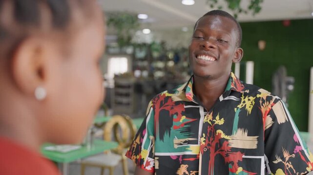 Black man and woman on date sharing shy smiles in bright modern cafe, young student man offering compliment with attentive gaze, colorful patterned shirt, playful laughter, subtle hand gesture