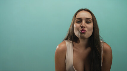 Naklejka premium Young woman puckers lips showing cleavage and bare shoulders in a teal studio wearing a beige tank top; flirty confidence.