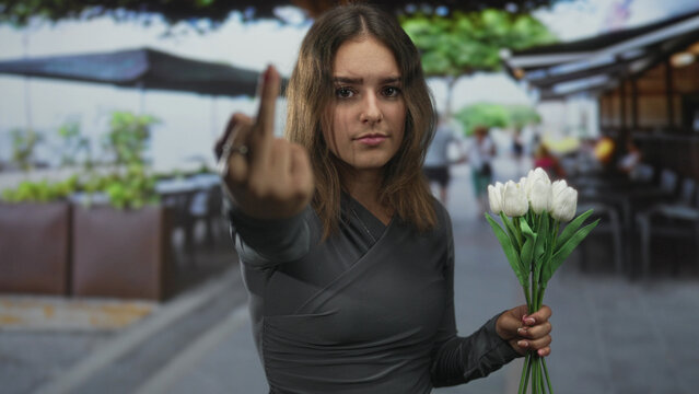 Young hispanic woman showing middle finger to camera while holding bouquet of white tulips on a restaurant terrace by the street; defiance.