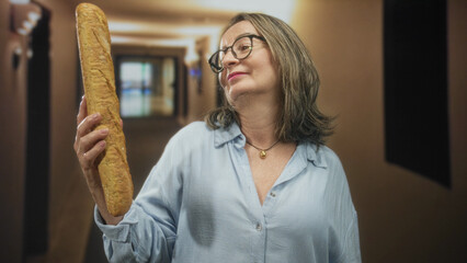 Hispanic senior woman holds a baguette upright with right hand in a building corridor, wearing glasses and a light blue shirt, looking at the loaf with a mild smile  quiet contentment. © Krakenimages.com