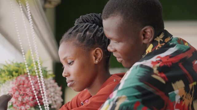 Black couple close embrace by window, sharing intimate home date with bouquet of flowers, soft sunlight illuminating braided hairstyle and colorful patterned shirt, quiet conversation and gentle