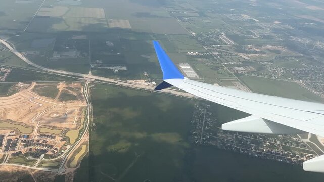 Airplane Wing View Through Window Over Rural America. Silver Plane with Blue Decor Flies to Cruising Altitude, Tilts from Farm Land to Clouds Travel