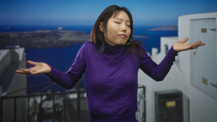 Obraz premium Woman in purple turtleneck raises both hands in a shrug gesture on a white seaside building terrace overlooking blue sea; quiet contemplation.