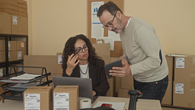 Man leans over showing a tablet to a seated woman at a packing desk with laptop, parcels, labels and scanner inside a building; concentration efficiency.