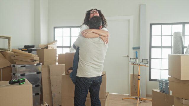 Man lifts woman in a tight hug amid stacked moving boxes, plastic bins, a work light and framed print in a building; moving day home joy.