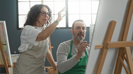 Obraz premium Woman instructor points finger at canvas while man paints on easel in art studio; creative mentorship focused.
