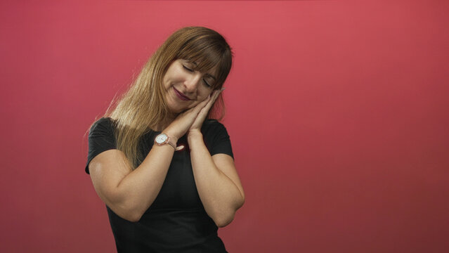 Young woman with hands to cheek in sleeping pose in studio with pink backdrop, wearing black t shirt and watch; calm rest.