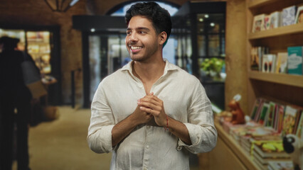 Obraz premium Man in linen shirt stands with hands behind back beside bookstore shelves inside a building; quiet contemplation.