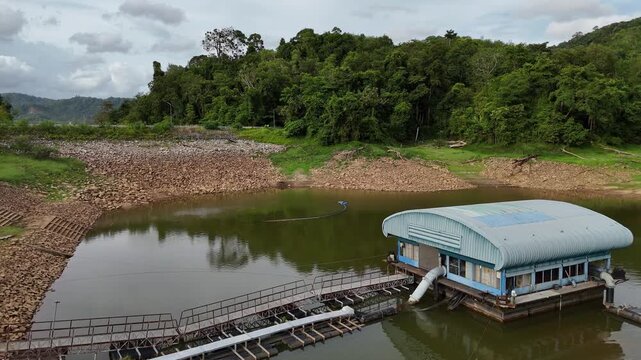 Aerial drone shot of water pumping station near dam spillway surrounded by tropical greenery and reservoir landscape. Industrial water management infrastructure with overflow pipe and calm lake scener