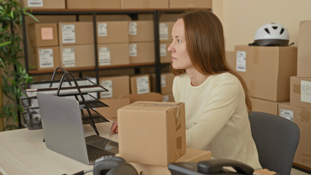 Woman at desk holds parcel with hand and checks laptop amid stacked boxes in a packing storage building; small business productivity focused.