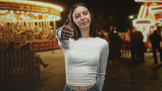 Young woman giving thumbsdown with extended thumb to camera at a nighttime carnival with carousel lights and blurred crowd; disapproval rejection.