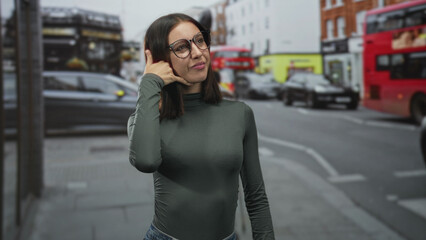 Fototapeta premium Woman wearing glasses makes a phone call gesture with thumb to ear and pinky to mouth while standing on a busy london street with traffic; playful confidence.