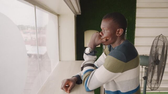 Black man smiling at window, leaning on counter with small fan beside him, morning light and striped sweater casual domestic scene, soft optimistic expression, planning surprise for partner, warm