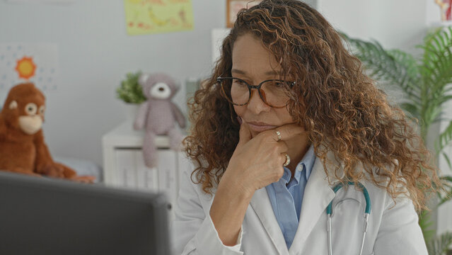 Woman doctor wearing glasses and white coat with stethoscope at computer, hand on chin, surrounded by stuffed toys and children's drawings in clinic building; focus.