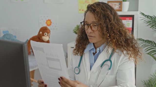 Woman doctor in lab coat with stethoscope reads printed report, fingers holding paper at computer in medical building; concentration.