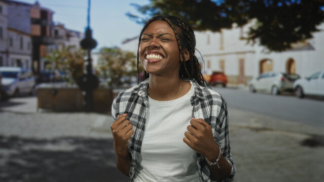 Woman with clenched fists pulling at her shirt while eyes closed and smiling on a sunny street; joy celebration.