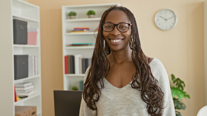 Woman wearing glasses looks at camera with neutral gaze and long braided hair, standing in a building study with bookshelves and clock  calm presence. © Krakenimages.com