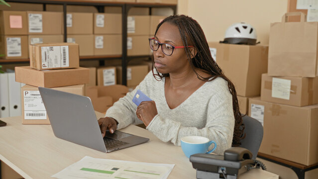 Woman holding creditcard and typing on laptop in a shipping area of a small business building with stacked parcels and mailing labels; determined small business owner.