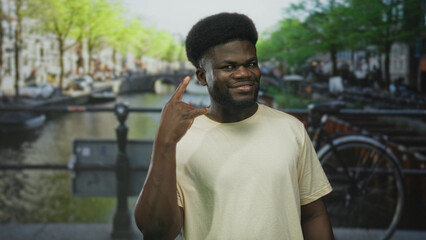 Fototapeta premium Man pointing finger to temple on street near canal and bicycle rack, smiling and looking at camera; playful confidence.