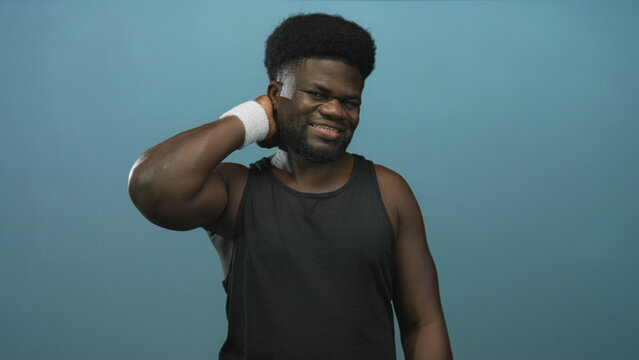 Man in black tank top with white wristband holds neck with right hand and winces against a blue studio wall; discomfort.