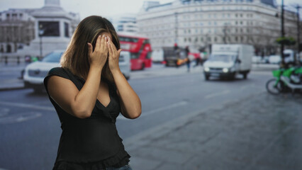 Fototapeta premium Woman covers face with hands on a busy city street with passing bus and van amid midday traffic and blurred buildings; vulnerability anxiety.