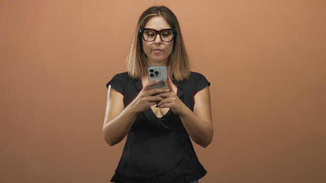 Woman wearing glasses holding smartphone with both hands, looking down and typing or scrolling on the device against a plain studio backdrop; quiet concentration.