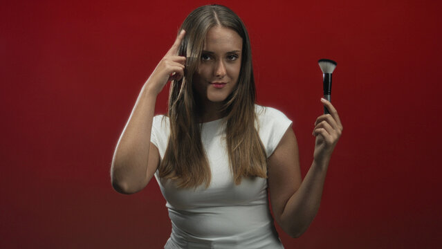 Woman in white top holds makeup brush and points finger to temple while smiling in studio; confidence beauty tutorial.