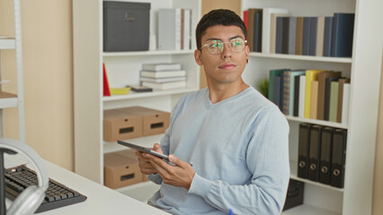 Fototapeta premium Man holding tablet, fingers tapping screen and looking up while seated at desk in a study building; focused study.