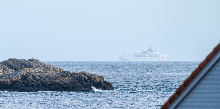 Lindesnes, Norway - April 04 2026: Ferry ship passing rocky shoreline seen between coastal house rooftops.