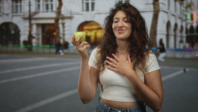 Young hispanic woman holding green apple on city street showing bare midriff, smiling and gesturing with hand near chest; joyful confidence.