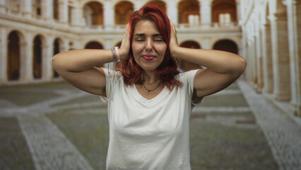 Fototapeta premium Woman with red hair covering ears with fingers in an old town stone building courtyard, eyes closed and smiling; playful defiance.