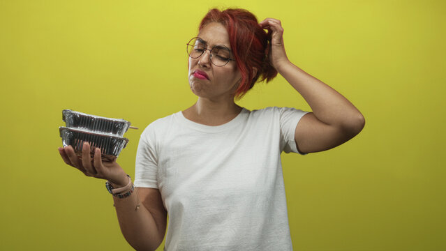 Young hispanic woman holding stacked foil takeout containers and scratching her head in a yellow studio; confusion.
