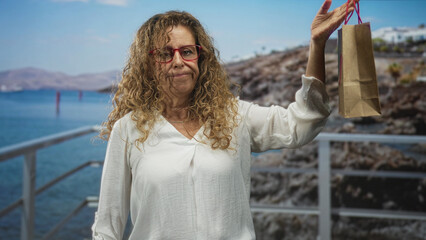 Fototapeta premium Woman in white blouse and red glasses holds small brown paper gift bag with heart aloft by right hand on seaside pier railing; gentle nostalgia.
