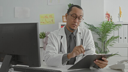Naklejka premium Man doctor in white coat holds pen and clipboard, points toward camera in clinic with stethoscope visible; telemedicine consultation reassurance.