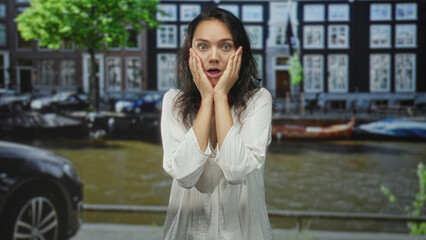 Fototapeta premium Woman in white blouse with hands pressed to cheeks showing surprised expression on a street canal with parked car and boat; surprise.