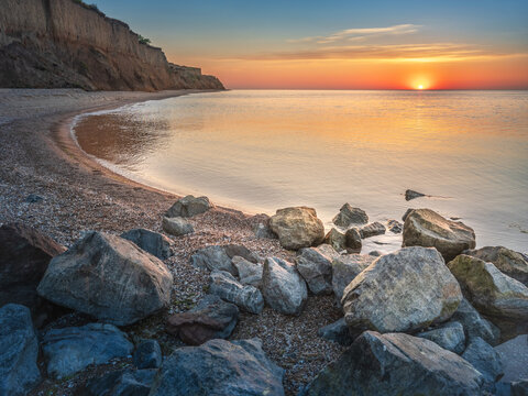 Curved sea coast with cliff and stones in water and rising sun