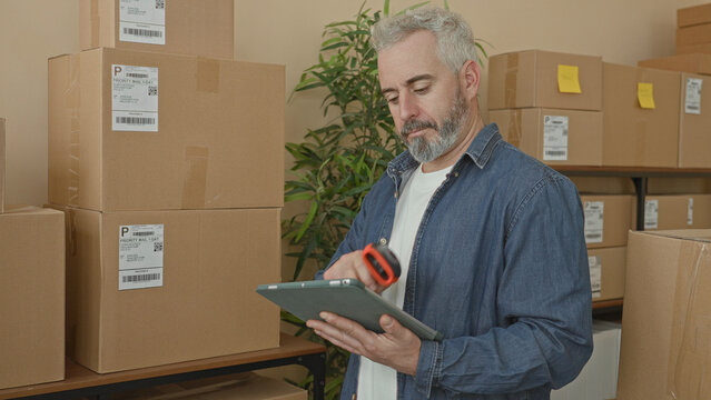 Man scans parcel with barcode scanner while holding tablet amid stacked boxes on shelf in warehouse; focused efficiency.