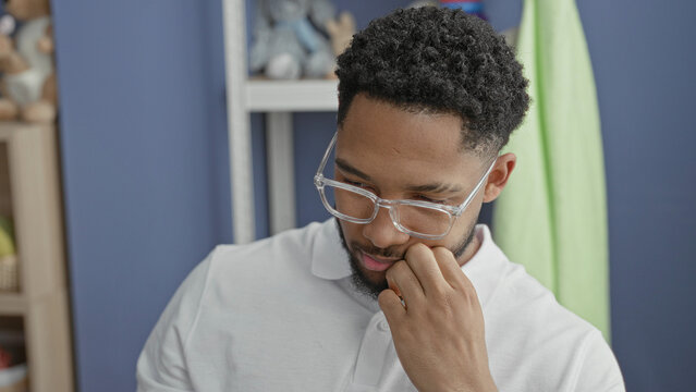 Man lifts clear glasses with one hand beside a baby crib in a nursery inside a building; fatigue quiet reflection.