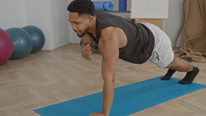 Fototapeta premium Man performing plank tap with one hand reaching on a blue mat, bare forearm visible in studio; focus determination.
