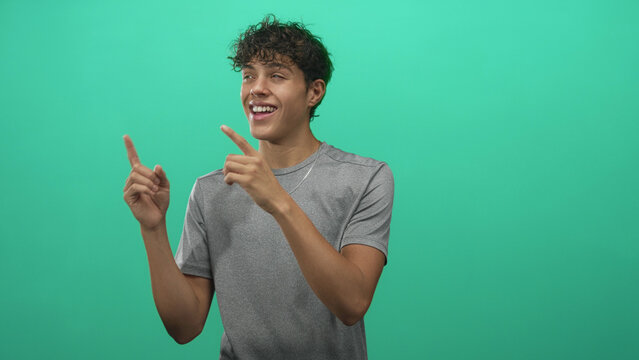 Teenage man wearing gray t shirt and chain necklace points both index fingers upward and smiles in studio; playful upbeat energy.