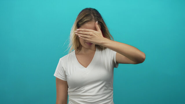 Woman covering her eyes with hand in studio against turquoise background, wearing white v neck t shirt, frontal pose; shy privacy.