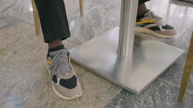 Sneakers under cafe table couple flirting with playful foot touch beneath metal table base on polished marble floor, casual trousers and patterned socks hinting at intimate daytime date, warm bistro
