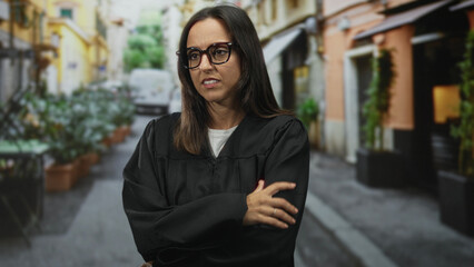 Fototapeta premium Woman judge in black robe wearing glasses with hand on upper arm and crossed posture in a narrow street; concern duty.