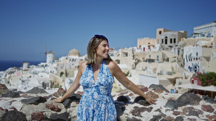 Fototapeta premium Woman leaning on stone wall building in santorini island town, hands on wall, smiling with sunglasses and blue floral dress; vacation joy.