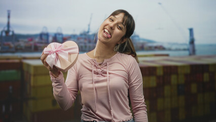 Fototapeta premium Young hispanic woman holding a heart shaped gift box, tongue out and smiling while presenting it at a shipping port with containers; playful romance.