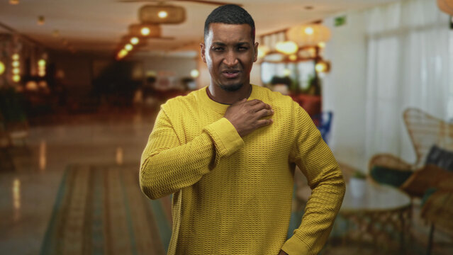 Man in yellow sweater clutching his shoulder with hand and wincing while standing in a hotel lobby reception area; pain health concern.