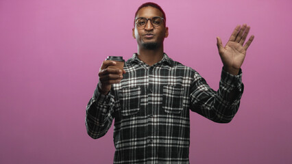 Naklejka premium Man wearing glasses and plaid shirt holding paper coffee cup and waving hand in studio with pink backdrop; friendly greeting.